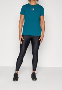 Person trägt ein türkisfarbenes Under Armour T-Shirt, schwarze glänzende Leggings und schwarze Sportschuhe und steht vor einem einfarbigen weißen Hintergrund.