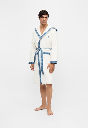 Homme debout pieds nus portant un peignoir blanc avec bordure bleue et capuche, attaché à la taille, sur un fond clair uni.