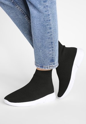 Füße, die schwarze sockenähnliche Sneaker mit weißen Sohlen und hochgekrempelten hellblauen Jeans vor einem schlichten weißen Hintergrund tragen.