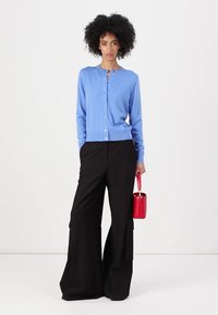 Cardigã azul claro com fecho de botão, emparelhado com calças largas pretas com bolsos; segurando uma pequena bolsa vermelha.