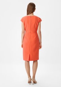 Robe à manches cap dans un tissu orange vif, silhouette ajustée, longueur genou, avec une fente au dos et une fermeture éclair discrète. Des talons beiges complètent le look.