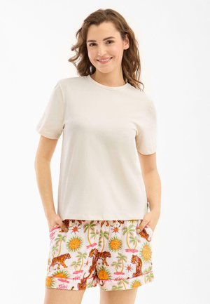 Jonge vrouw lacht, draagt een effen beige t-shirt en kleurrijke shorts met tijger-, palmboom- en zonpatronen, handen in de zakken.