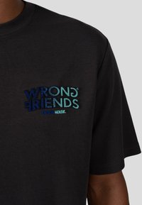 Černé bavlněné tričko s vícebarevným vyšitým textem: "WRONG FRIENDS DESIGN HOUSE," s různými odstíny modré a zelené.