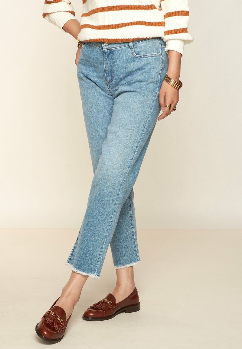 Hellblaue, hochtaillierte Jeans mit geradem Bein und fransigem Saum. Dazu dunkelbraune Slipper mit Fransen und goldenen Akzenten.