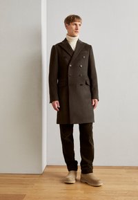 Abrigo de lana de doble botonadura en verde oscuro con seis botones, bolsillos laterales, solapas en pico, combinado con pantalones verde oliva y botas beige.