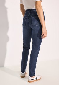 Jeans ajustados de color azul oscuro con una cintura alta, que presentan bolsillos traseros y una textura de denim suave. Combinados con zapatillas blancas, de pie contra un fondo claro.