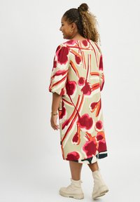Kleid mit Blumenmuster in Creme mit roten und orangenen Motiven. Hat Puffärmel, einen lockeren Schnitt und reicht bis unter das Knie. Getragen mit beigen Stiefeln.