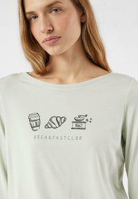 Jasnozielona koszula z długim rękawem z graficznymi ilustracjami filiżanki kawy, rogalika i młynka oraz napisem "BREAKFASTCLUB".