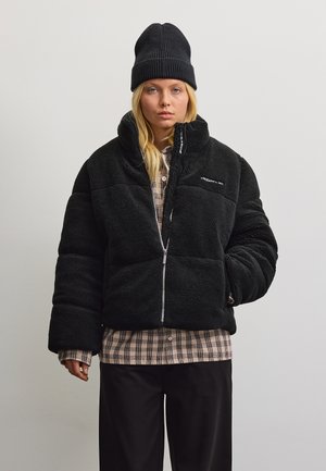 Jeune femme portant un bonnet noir, une doudoune noire, une chemise à carreaux et un pantalon noir, debout devant un fond clair uni.