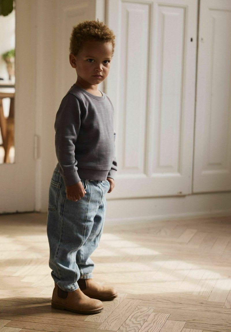 Jeune garçon aux cheveux bouclés portant un sweat-shirt gris, un jean ample et des bottes marron, debout à l'intérieur sur un sol en bois près de portes blanches.