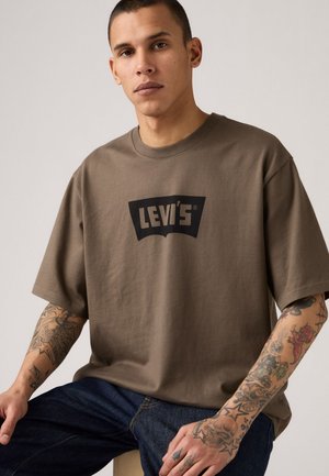 Brown cotton t-shirt with a large black Levi's logo on the front, featuring a relaxed fit and short sleeves. Denim jeans visible below.