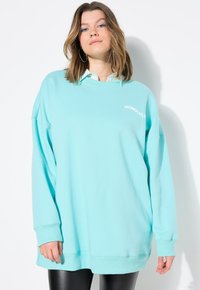 Sudadera de gran tamaño en azul claro con puños y dobladillo acanalados, con el texto "HONOLULU" en blanco. Lleva una camisa de rayas con cuello debajo.
