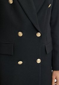 Svart skräddarsydd kappa med dubbelknäppt design, texturerad tyg och utsmyckade guldknappar med ett blommönster.