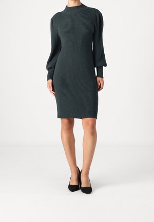 Frau trägt ein dunkelgrünes, geripptes Strickkleid mit langen, aufgebauschten Ärmeln und schwarzen High Heels, steht vor einem schlichten weißen Hintergrund.