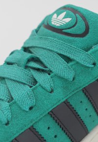 Zbliżenie na turkusowy zamszowy but sportowy Adidas z teksturowanymi sznurowadłami, czarnymi paskami i białym logo Trefoil na języku.