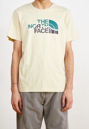 Mężczyzna ubrany w jasnżółty krótki T-shirt z turkusowo-czarnym logo "THE NORTH FACE" oraz szare spodnie.