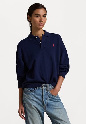 Mørkeblå polo skjorte med korte ermer og krage, med en rød logo på brystet. Kombinert med lyseblå denim jeans.