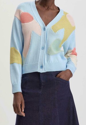 Femme portant un cardigan bleu clair boutonné avec des taches pastel abstraites et une jupe bleu foncé, une main dans la poche.