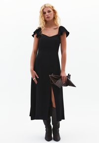Vestido maxi negro con mangas cortas con volantes, escote cuadrado y una abertura frontal. Llevado con botas marrones hasta la rodilla y sosteniendo un clutch marrón.