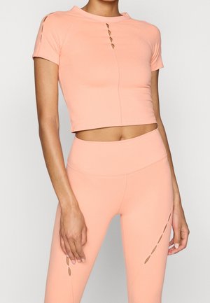 Kvinde iført ferskenfarvet crop top med korte ærmer og højtaljede leggings med udskæringer på brystet, benet og ærmet.