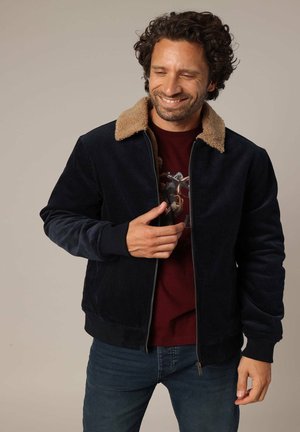Veste en velours côtelé bleu marine avec un col en fausse fourrure beige, des poignets et une taille côtelés. Fermeture éclair à l'avant, portée sur un t-shirt graphique bordeaux.
