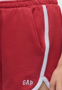 Κόκκινα αθλητικά σορτσάκια με λευκή επένδυση και λογότυπο GAP, με ελαστική μέση και πλαϊνή τσέπη, φορεμένα στη λεκάνη ενός ατόμου με ορατό χέρι.