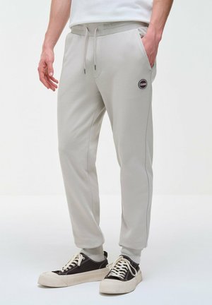 Pantaloni da jogging grigio chiaro con polsini elasticizzati e coulisse in vita, patch con logo sulla coscia, indossati con scarpe da ginnastica bianche e nere.