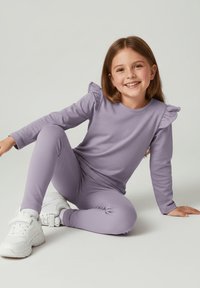 Lawendowy zestaw do noszenia w domu, składający się z długiego topu z falbankami i dopasowanych spodni. Noszony z białymi sportowymi sneakersami.