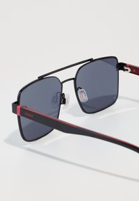 Ochelari de soare pătrați cu rame negre, lentile gri și accente roșii pe tâmple. Dotati cu pernițe ajustabile pentru confort. Branding HUGO vizibil.