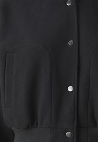 Svart jacka med en mjuk tygstruktur, tryckknappar, stora fickor och en åtsittande midja för en strukturerad look.