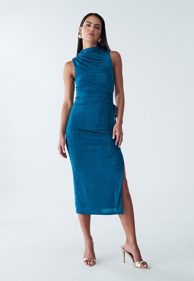 Vestido midi en color azul verdoso, confeccionado en una tela suave y elástica, con un cuello alto, detalle drapeado, abertura lateral y un acento de lazo. Combina con sandalias de tacón doradas.