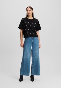 Schwarzes Baumwoll-T-Shirt mit kurzen Ärmeln und weißen Grafikdrucks, kombiniert mit hoch sitzenden blauen weiten Jeans und schwarzen offenen Schuhen.