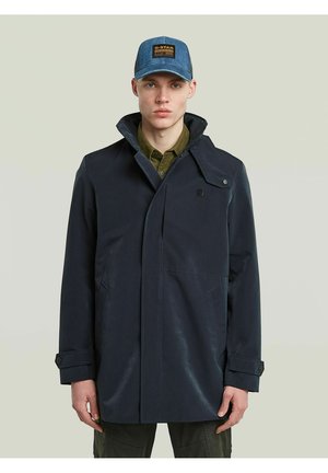 Junger Mann trägt einen dunkelblauen Mantel über einem olivfarbenen Hemd und eine blaue Jeanskappe mit braunem Logo-Patch, stehend vor einem einfarbigen Hintergrund.