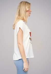 T-shirt blanche à manches courtes avec une coupe décontractée, arborant un motif floral rouge sur le devant. Associée à un jean bleu clair. Tissu en coton doux.