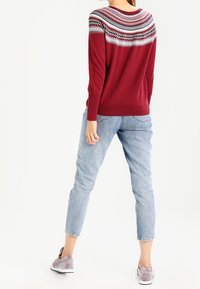 Femme debout de dos portant un pull en maille rouge à motif, un jean bleu clair et des baskets grises sur fond blanc.