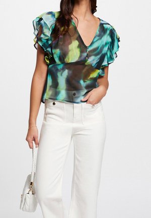 Vrouw draagt een gerimpelde, meerkleurige blauw-groene blouse met witte hoge taille broek, houdt een witte tas met een gevlochten handvat vast.