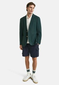 Blazer vert avec boutons dorés, polo blanc, shorts cargo bleu marine, baskets blanches et chaussettes rayées. Design simple avec un look superposé.