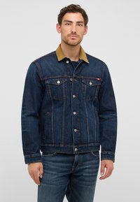 Homme portant une veste en denim bleu foncé avec un col en velours côtelé beige et un jean bleu assorti, se tenant contre un fond gris clair uni.