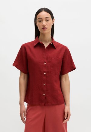 Femme aux cheveux raides et foncés portant une chemise rouge à manches courtes boutonnée avec col et un pantalon rouge assorti sur un fond uni.