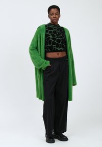 Grüner, flauschiger langer Cardigan, schwarzer, taillierter Pullover mit Giraffenmuster, lockere schwarze Hose und schwarze Ankle Boots. Glatte Textur im gesamten Outfit.