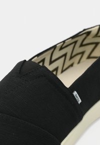 Chaussure slip-on en toile noire avec une doublure intérieure à motif zigzag et une petite étiquette en tissu blanc portant l’inscription "TOMS" sur le côté.