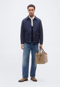 Άνδρας που φοράει ναυτικό μπουφάν, λευκό πουκάμισο, μπλε τζιν και καφέ μπότες, κρατώντας μια ταμπά τσάντα duffel από καστόρι και δέρμα πάνω σε λευκό φόντο.