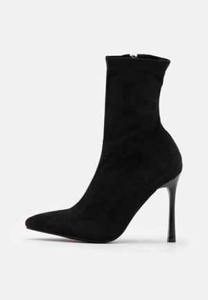 Zwarte suède enkellaars met spits toelopende neus, slanke hoge naaldhak en ritssluiting aan de zijkant, weergegeven op een witte achtergrond.
