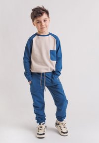 Młody chłopiec stoi z rękami w kieszeniach, ubrany w niebiesko-beżową bluzę, pasujące niebieskie spodnie dresowe i białe trampki na białym tle.