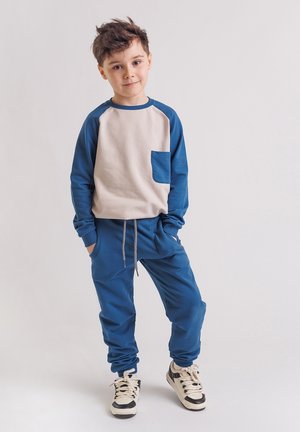 Młody chłopiec stoi z rękami w kieszeniach, ubrany w niebiesko-beżową bluzę, pasujące niebieskie spodnie dresowe i białe trampki na białym tle.