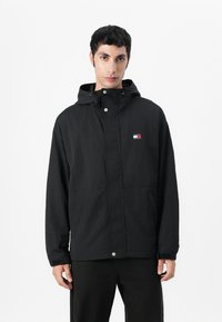 Zwarte hoodie-jack met een rits- en drukknoopsluiting, gestructureerde stof, zijzakken en een klein logopatch met rode, witte en blauwe accenten.