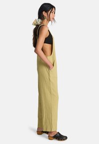 Λινοφόρεμα ανοιχτού πράσινου χρώματος με υφή, πλαϊνές τσέπες και ανοιχτές πλευρές, σε συνδυασμό με μαύρο κοντό τοπ και μαύρες ξύλινες παντόφλες.