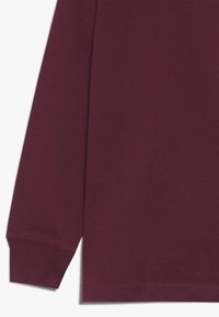 Burgundy långärmad skjorta med en åtsittande manschett och slät bomullstextur. Har en rak fåll och enkel design utan mönster eller detaljer.