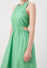 Vestido verde sin mangas con corsé ajustado, cinturilla elástica y detalles recortados; hecho de material ligero con una textura suave.