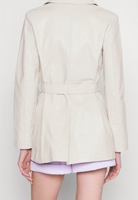 Veste en cuir beige clair avec une taille ceinturée, une coupe ajustée et des manches longues. Le dos présente une fente et une texture lisse. Short violet visible en dessous.